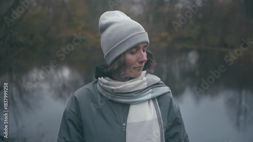 Cold lake A beautiful Slavic woman in a cozy grey jacket and checkered scarf gazes thoughtfully by a serene lake surrounded by tranquil autumn colors.
