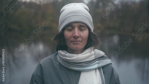 Enjoying nature: A beautiful Slavic girl dressed in a grey jacket and beanie stands by a tranquil lake, lost in reflection amidst a serene autumn landscape with soft, muted colors.