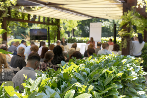 Outdoor business conference with professional audience attending presentation in green garden venue under canopy, sustainable corporate event setting