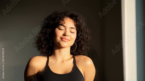 Young woman enjoying sunlight with closed eye serene curly hair hoop earring tank top warm glow relaxing sunlight peaceful expression stretching neck