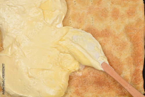 A close-up, high-angle view of a wooden spoon spreading creamy yellow custard filling over a sheet of baked puff pastry on a dark baking tray