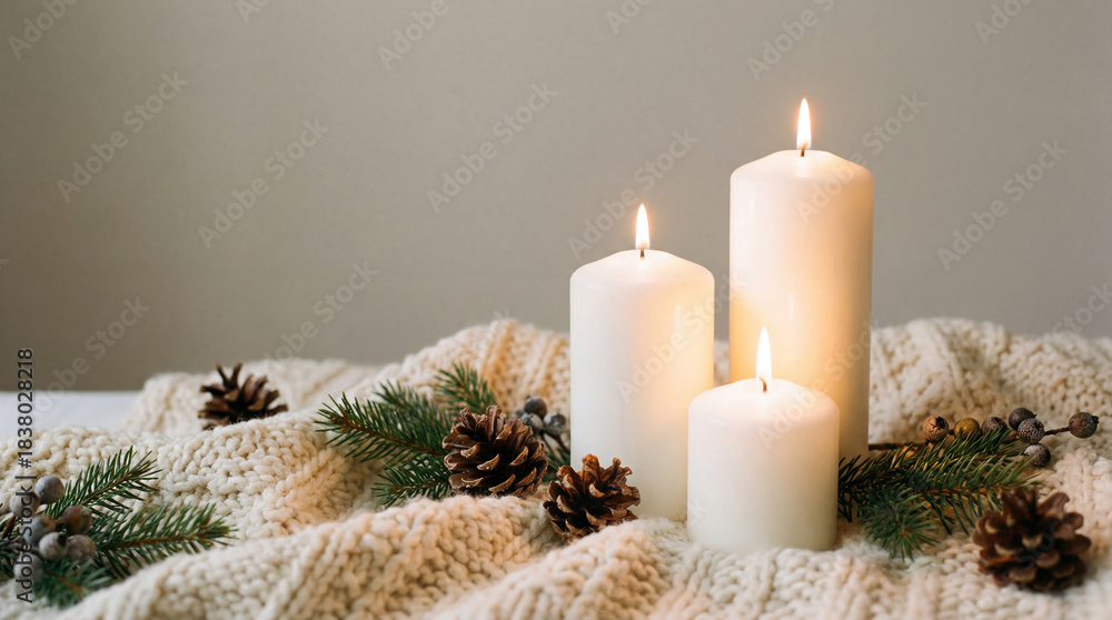 Naklejka premium Cozy winter arrangement with three white candles on knitted blanket decorated with pinecones and evergreen branches indoors