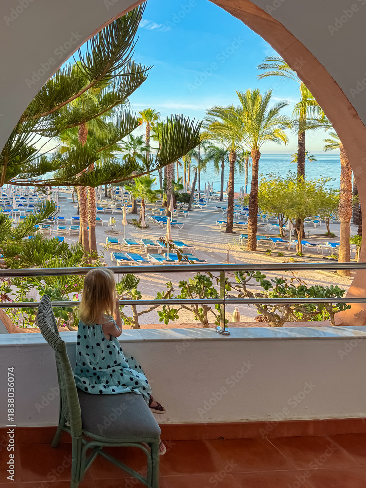 Obraz premium A little girl sits on a balcony chair gazing at the sandy beach and palm trees of a luxury resort in Almunecar. The child enjoys the ocean view through the terrace railing during a sunny holiday.