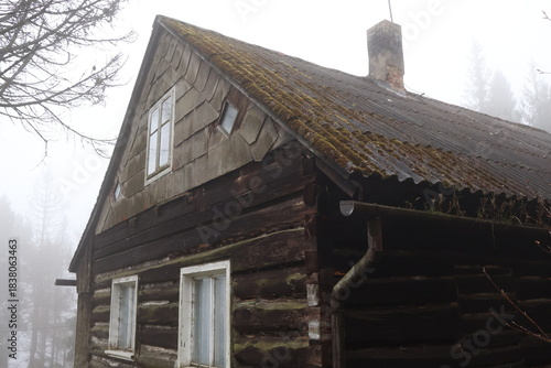 Fotografía Old wooden log cabin with mossy roof tiles and brick chimney in heavy fog