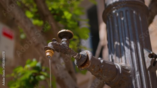 A person hand is captured mid-action turning a decorative vintage-style outdoor water tap attached to a cast iron fountain with blurred green foliage in the background