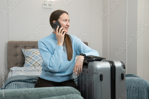 Young woman with long brown hair, wearing a light blue sweater, sitting on a bed, leaning on a gray suitcase, talking on her mobile phone while planning travel arrangements