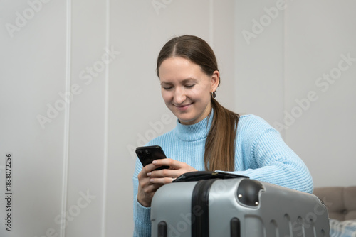Young woman waiting for a flight at home, using her smartphone to make travel arrangements while her passport and suitcase sit nearby, radiating excitement for the upcoming trip