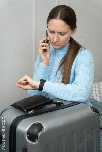 Young businesswoman sitting on bed with suitcase, looking at smartwatch and making phone call, possibly arranging transportation or confirming arrival time. Vertical photo