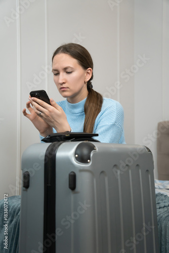 Young woman sitting on bed next to a packed suitcase, using her smartphone for travel preparations, planning her upcoming trip and managing online bookings for her journey. Vertical photo