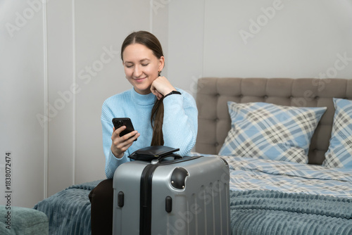 Young woman sitting on a bed next to a packed suitcase, using her smartphone to finalize travel plans for an upcoming business trip or vacation, embracing modern technology for convenience