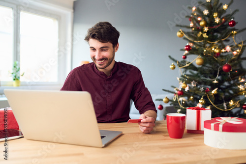 Smiling young man doing Christmas online shopping with laptop paying with credit card