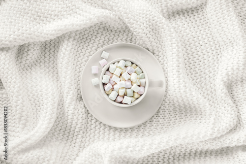 Cup with pastel marshmallows on soft white knitted blanket. Cozy winter still life, top view, flat lay. Color of the year 2026 - Cloud Dancer