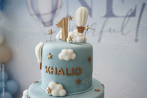 A soft blue two-tier first birthday cake decorated with clouds, hot air balloons, and gold accents. A stylish and whimsical cake design perfect for baby celebrations, party styling, and milestone even