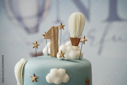 Close-up detail of a pastel blue first birthday cake featuring a gold number one, hot air balloon topper, fluffy cloud decorations, and star accents. A soft, elegant design perfect for baby celebratio
