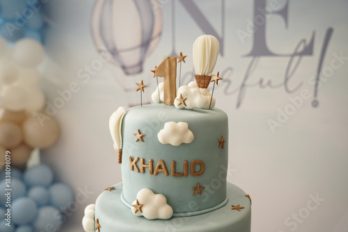 A soft blue two-tier first birthday cake decorated with clouds, hot air balloons, and gold accents. A stylish and whimsical cake design perfect for baby celebrations, party styling, and milestone even