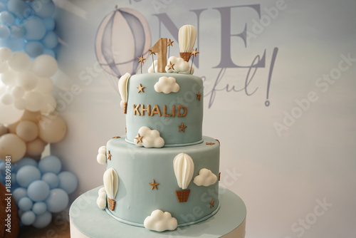 A soft blue two-tier first birthday cake decorated with clouds, hot air balloons, and gold accents. A stylish and whimsical cake design perfect for baby celebrations, party styling, and milestone even