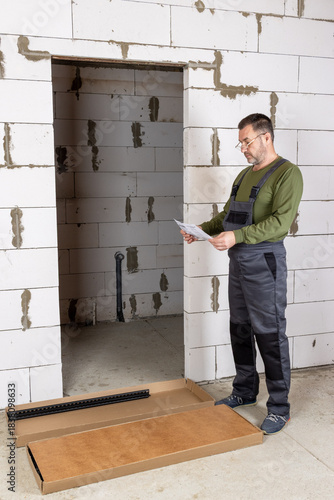 A middle-aged man in glasses intently studies the assembly manual for a metal shelving unit in an unrenovated room