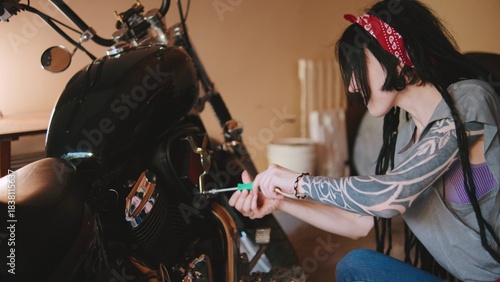 Woman repairing motorcycle in garage while wearing bandana and demonstrating mechanical skill with tools