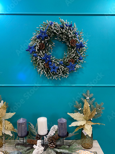 Christmas wreath with blue glitter stars and pinecones hanging on a teal wall, with a table centerpiece featuring candles, frosted greenery, and gold poinsettia flowers