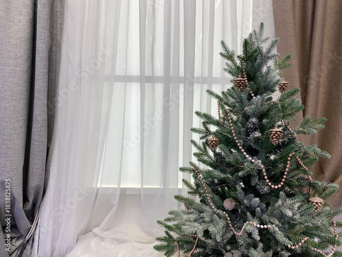 christmas tree in the window. A modern white matte Christmas tree topped with rose gold garland and glittery pine cones sits against a window with sheer white curtains and taupe drapes.