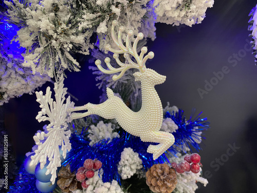 Close-up of a Christmas tree branch with a white beaded reindeer ornament, snow-covered pine needles, blue tinsel, red berries, and a large snowflake, all under blue lighting