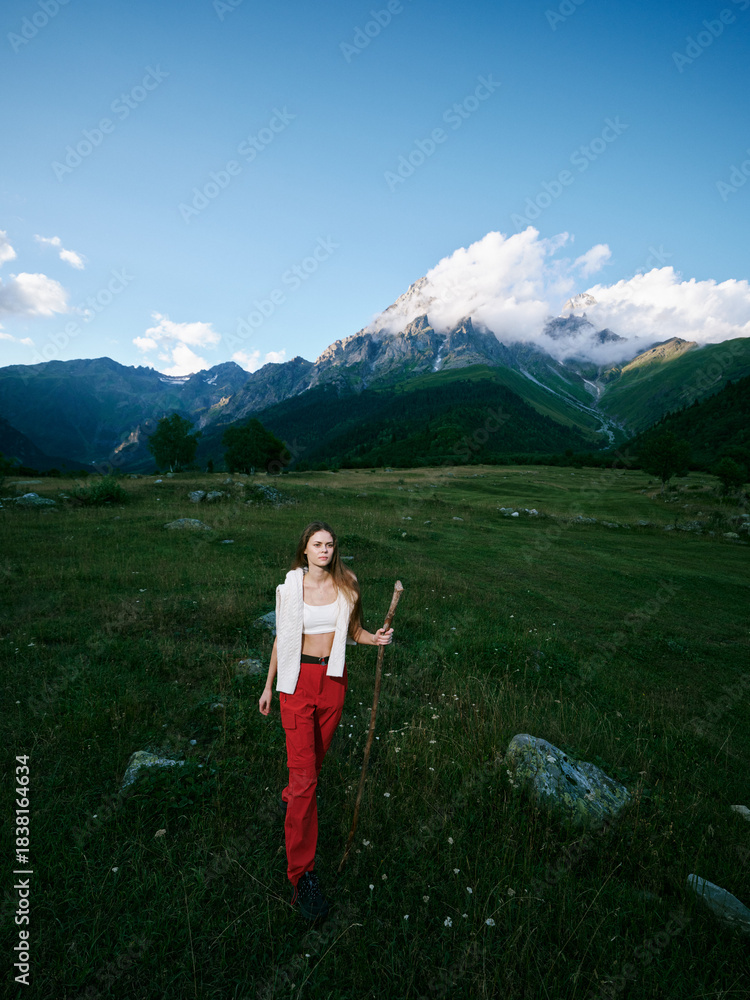 Fototapeta premium Mountain landscape, outdoor scene with a woman wearing red pants standing in a meadow, distant peaks, clear sky, rugged terrain and calm atmosphere, ideal for travel, exploration and nature