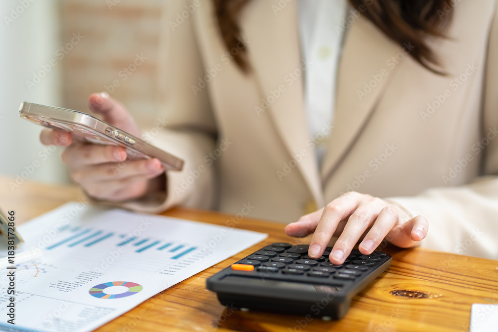 Fototapeta premium Businesswoman using smartphone and calculator for financial analysis in office professional environment close-up view
