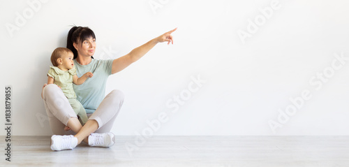 Fotografie Mother sitting with her baby on floor and pointing aside at copy space, guiding the child attention