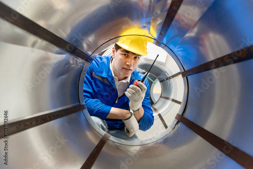 Male worker with safety clothing and helmet holding walkie talkie during inspecting large metal tube in industrial site. Engineer work at manufacturing factory. Inspector checking quality of product.