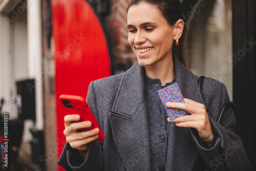Stylish woman holding smartphone and credit card while making a mobile payment outdoors. Cashless payment, fintech, digital wallet and modern urban lifestyle concept. Happy smiling woman in grey coat.