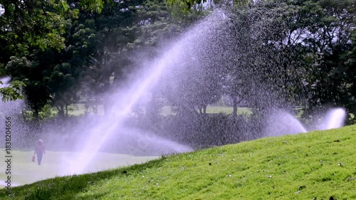 High-pressure sprinklers watering the pristine green lawn of a golf course, featuring a sand bunker edge in the lower foreground.