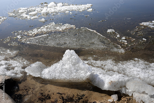 Unique ice formations are visible on the water surface, contrasting with the sandy shore. The scene captures the beauty of nature, showcasing textures and reflections in a serene environment