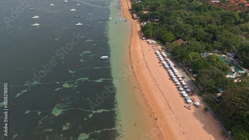 High angle orbit view of sandy beach with parasols, boats, and clear water in Sanur