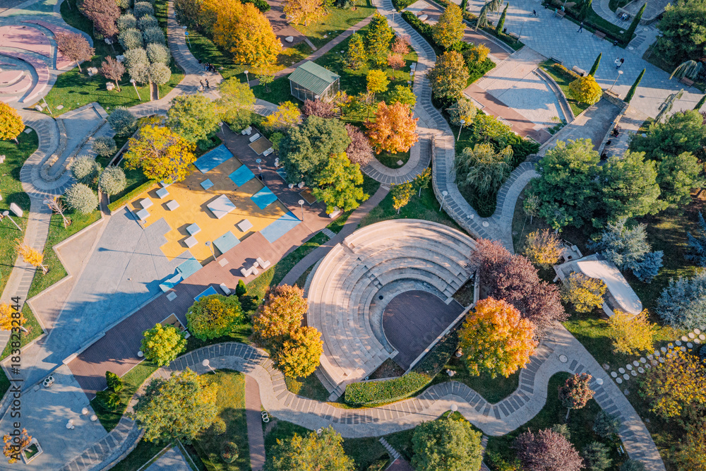 Naklejka premium Urban park featuring an open air amphitheater, winding pathways, colorful trees, and recreational areas from an aerial perspective during autumn