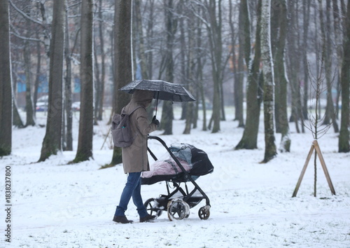 A young mother with a baby in a stroller walks through a snow-covered park in a snowstorm under an umbrella, Yesenin Park, Saint Petersburg, Russia, December 08, 2025