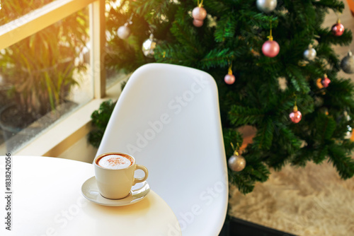 Embracing holiday spirit with delicious hot cappuccino on table near window and beautiful view of decorated Christmas tree in background. hot cup of coffee with Merry Christmas festive decorations.