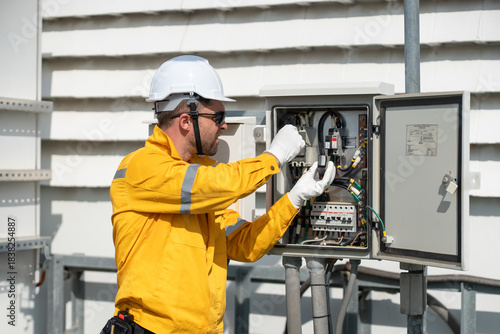 Engineer in safety helmets and yellow jackets inspecting electrical control cabinet with laptop and tablet on site. Mobile phone Telecommunication Radio antenna tower installed on building