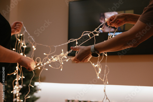 Hands arranging warm white Christmas lights for holiday decorating. Two people carefully handle a string of festive lights, preparing for the cozy winter holiday season. New Year 2026 home party.