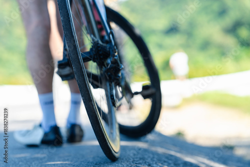 A close-up of a bicycle wheel on a road, with a cyclist's legs visible, set against a blurred green background.
