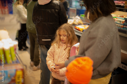 Young girl at supermarket checkout, unspecified race, awaiting purchase with parent beside register. Pink jacket child stands near orangehatted