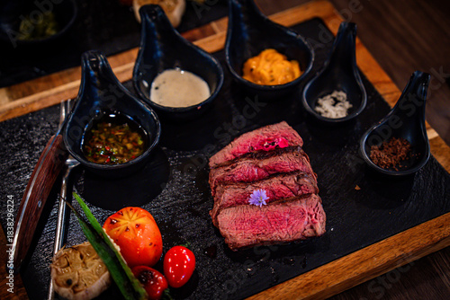 A gourmet grilled steak served on a rustic black slate board with assorted sauces, roasted garlic, cherry tomatoes, and okra. The rich texture and elegant plating highlight a premium dining experience