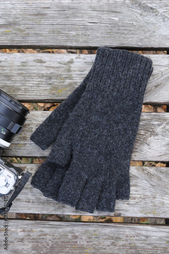 Photo camera and gloves with open fingers on wooden background. Warm knitted handmade things for photographers.