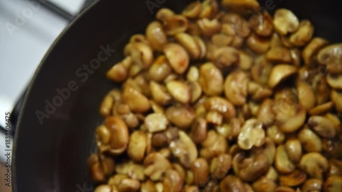 A slow-motion close-up of sliced small porcini mushrooms being fried in a frying pan in a home kitchen. Stirring the mushrooms as they fry by tossing them in the pan.