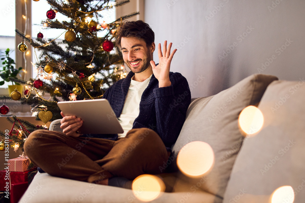 Obraz premium Smiling young man using digital tablet for video call sitting on sofa during Christmas season
