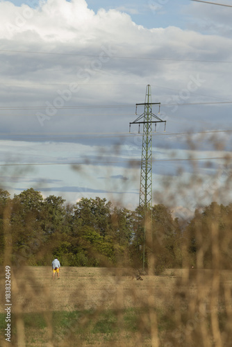 white man walking dog under pylon, golden dry grass in foreground, distant tree line, layered clouds above, late afternoon light,