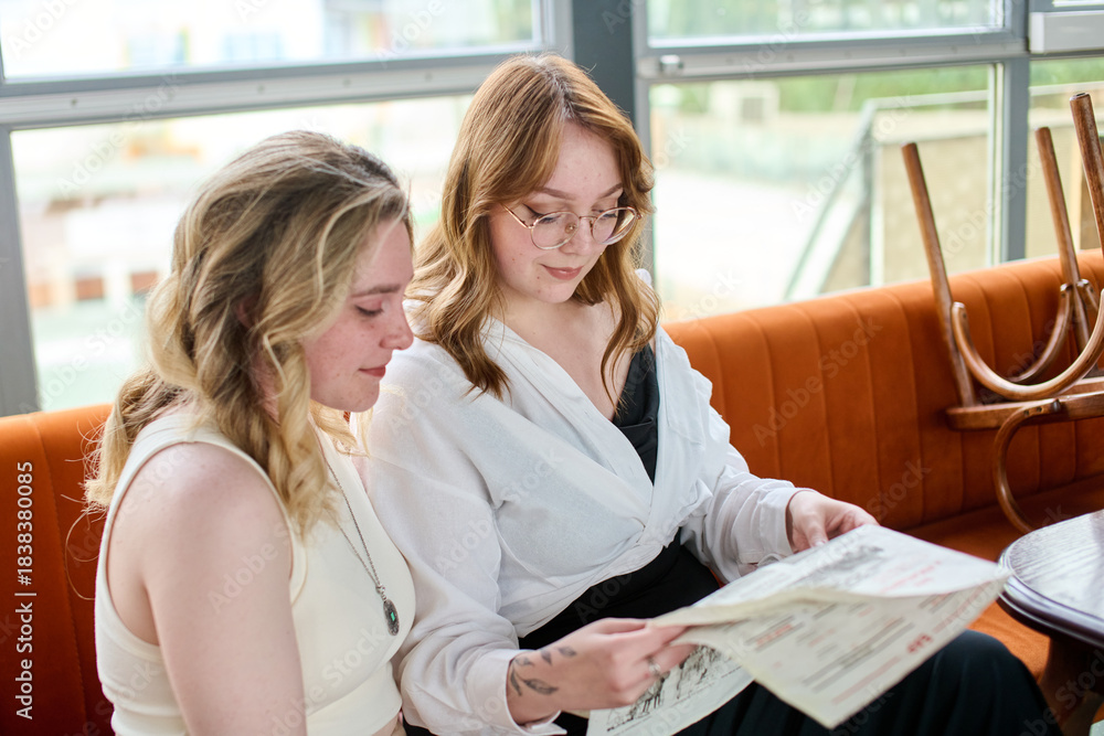 © TRAVELARIUM - Two young women reading menu in bright cafe with large windows