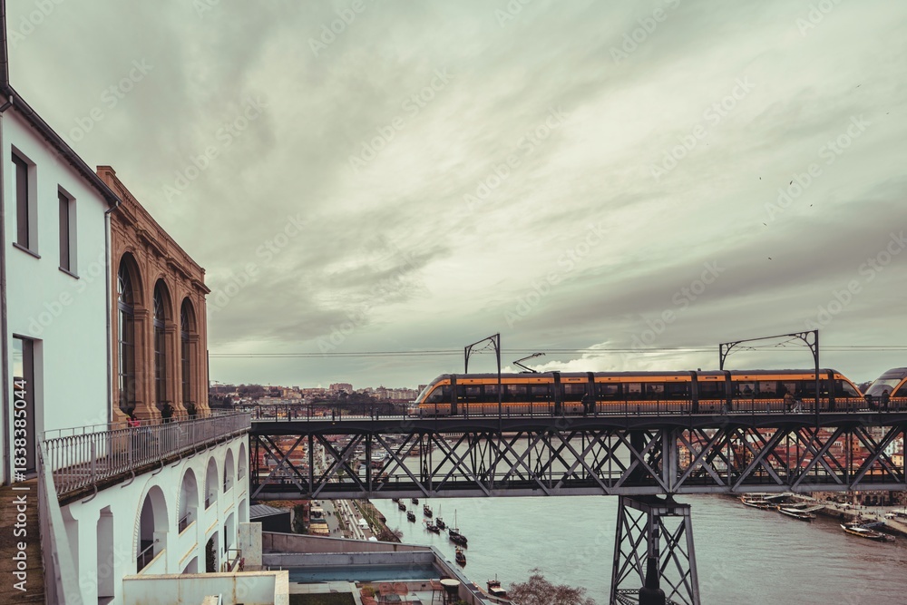 Obraz premium A vivid yellow train traverses a metallic bridge over the Douro River. The sky is filled with dramatic clouds, while a classic building stands adjacent