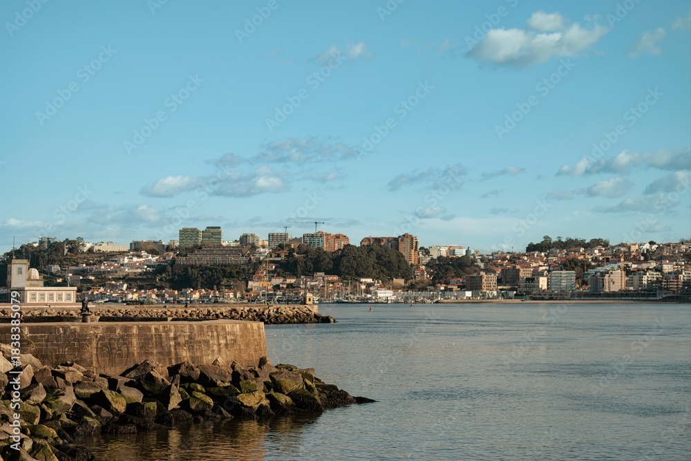 Obraz premium Buildings on a hillside are across a calm river. Rocks are piled up on the shoreline. A peaceful and serene scene unfolds in Vila Nova de Gaia