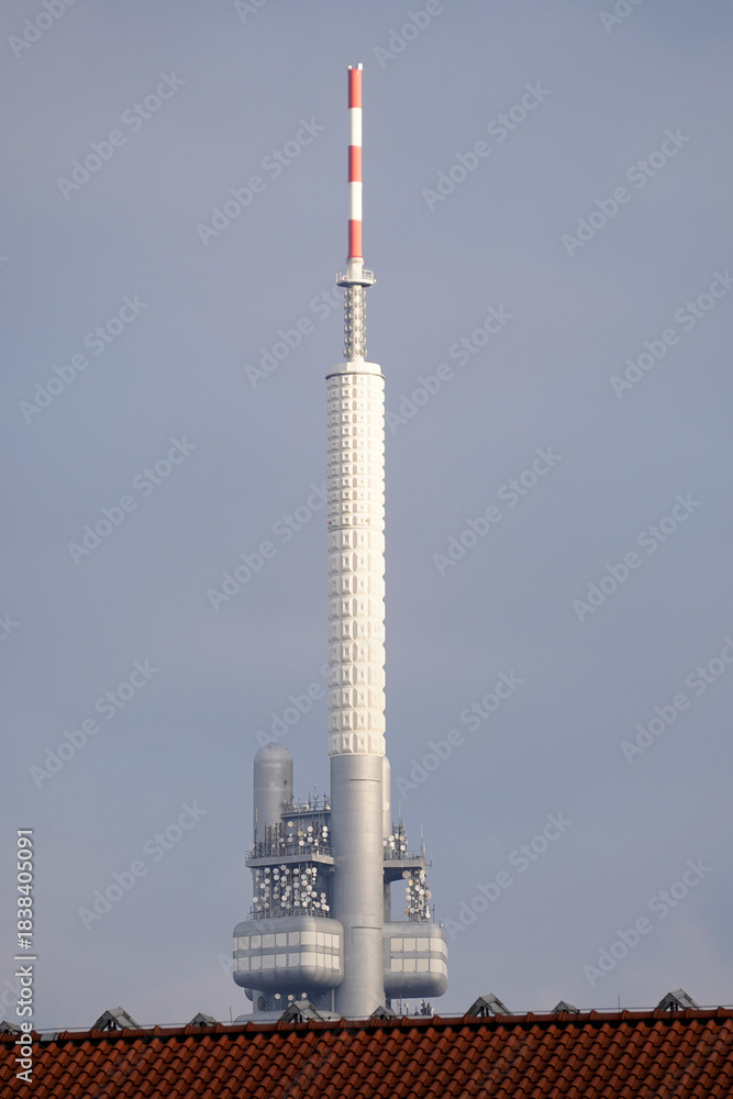 Obraz premium Tall telecommunications tower with antennas rising above rooftops against a clear sky. Broadcasting infrastructure, signal transmission technology, vertical structure, and urban communication network.