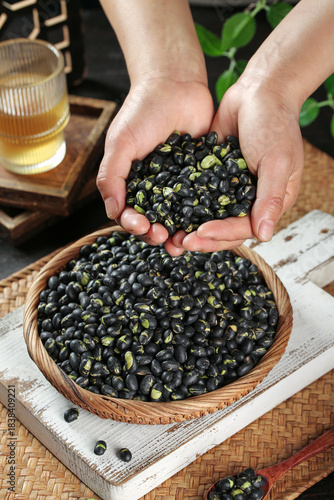 Fotografie Fresh Black Beans in Hands from Wicker Basket on Rustic Wood Table